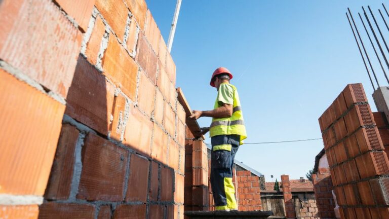 Trabalhador da construção civil erguendo paredes de tijolos, etapa que compõe o memorial descritivo do imóvel.